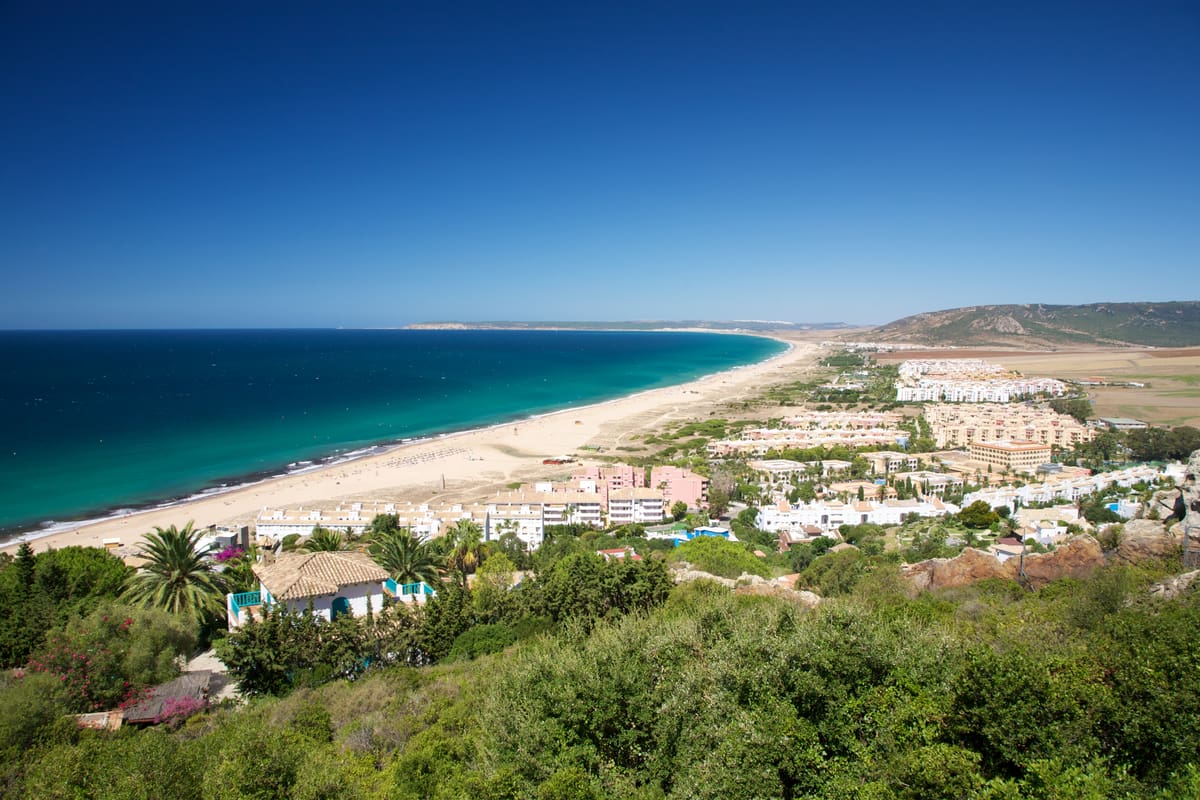 A panoramic view of a coastal town with white buildings, green vegetation, a long sandy beach, and turquoise blue sea under a clear sky. Hills are visible in the background. Zahara de los Atunes, Cadiz, Costa de la Luz