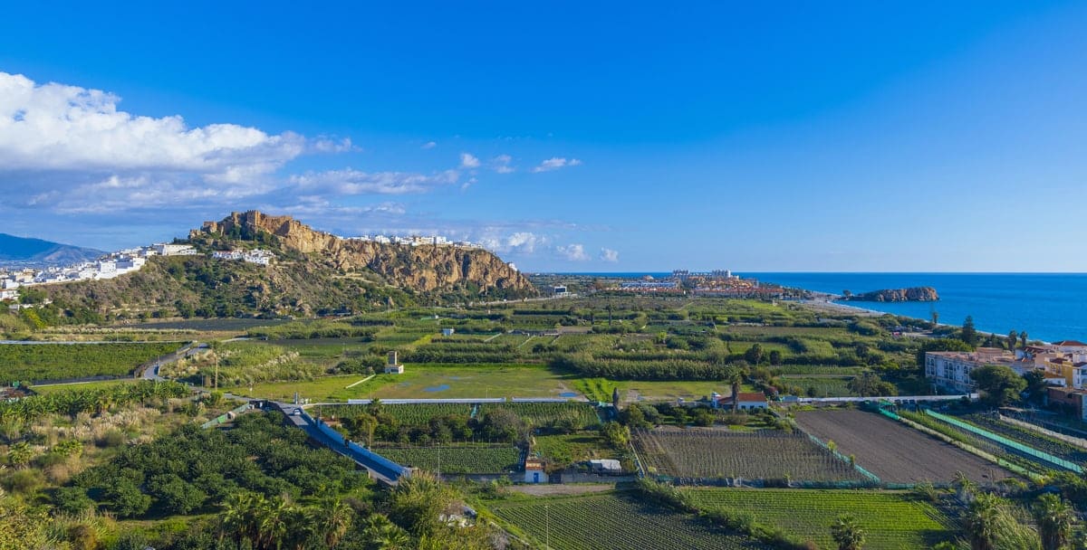 A coastal landscape with green fields, a rocky hill with white buildings on top, and the blue sea stretching to the right under a clear sky. Costa Tropical with Salobreña