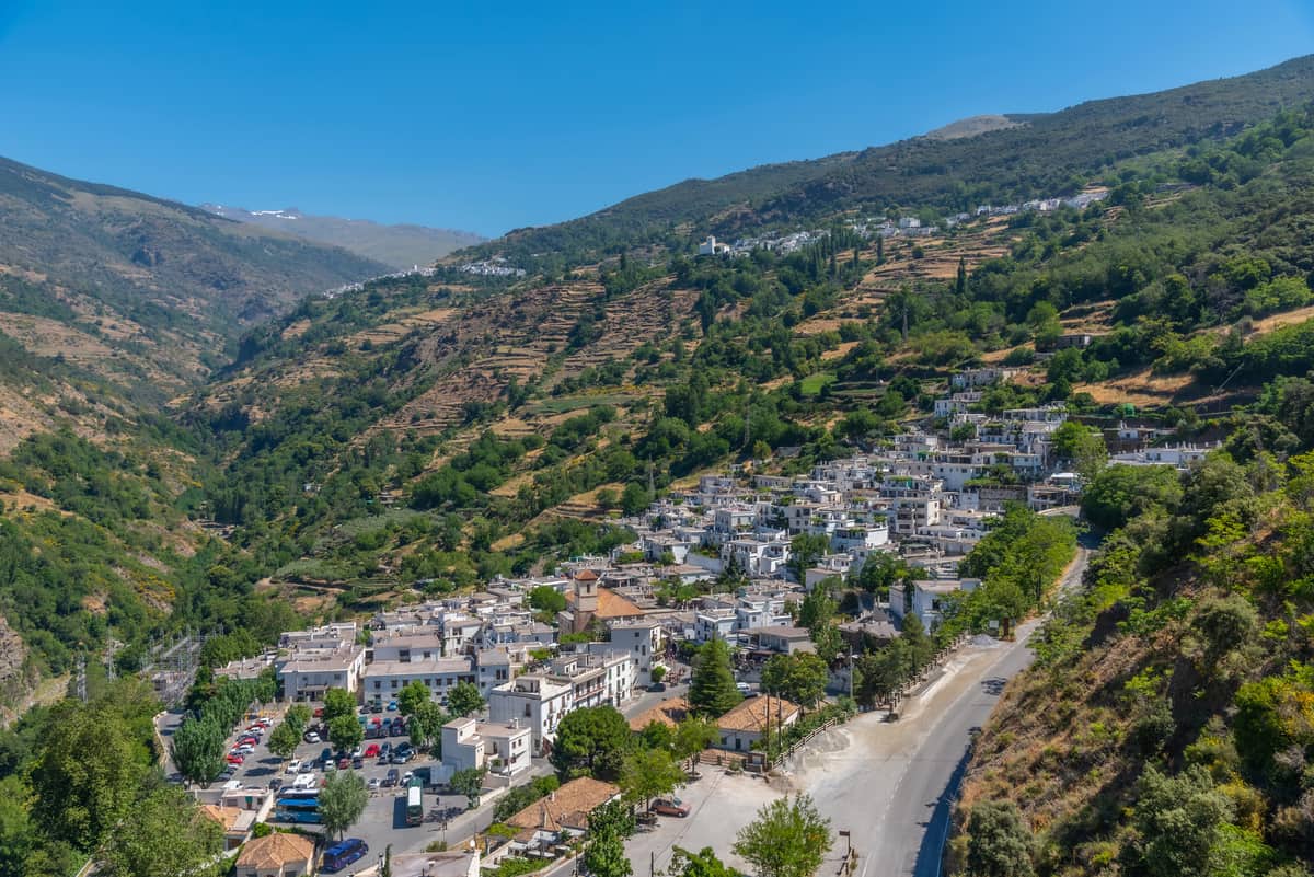 A small white village sits on a green hillside surrounded by mountains, with terraced farmland above and a winding road below under a clear blue sky. Pampaneira in Las Alpujarras