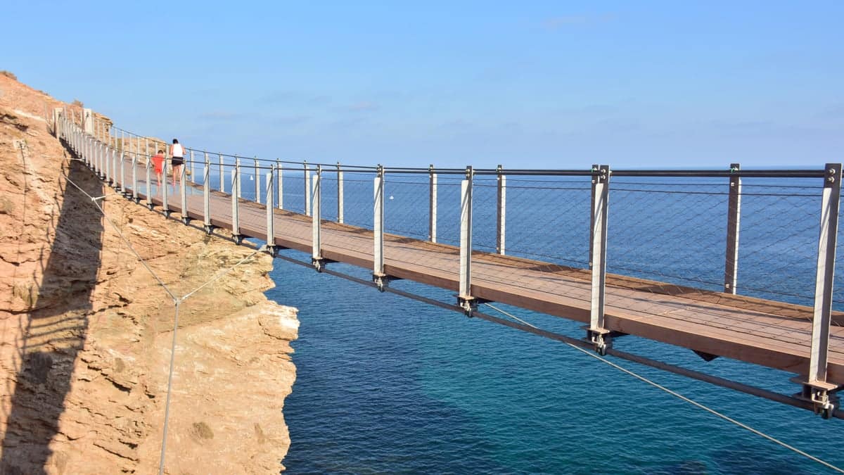A person walks across a narrow suspension bridge built along a rocky cliff above clear blue sea water under a bright sky. Pasarela Colgante de Jolúcar in Torrenueva Costa