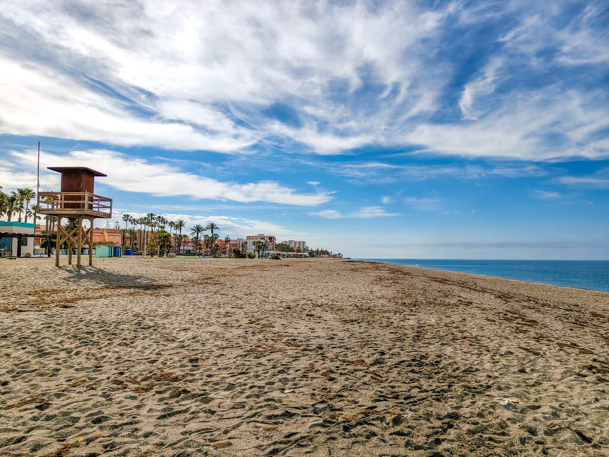 A wide sandy beach with a wooden lifeguard tower, palm trees, buildings in the distance, and a blue sky with scattered clouds. The sea is calm along the right edge of the image. Playa de la Charca-Salomar in Salobreña