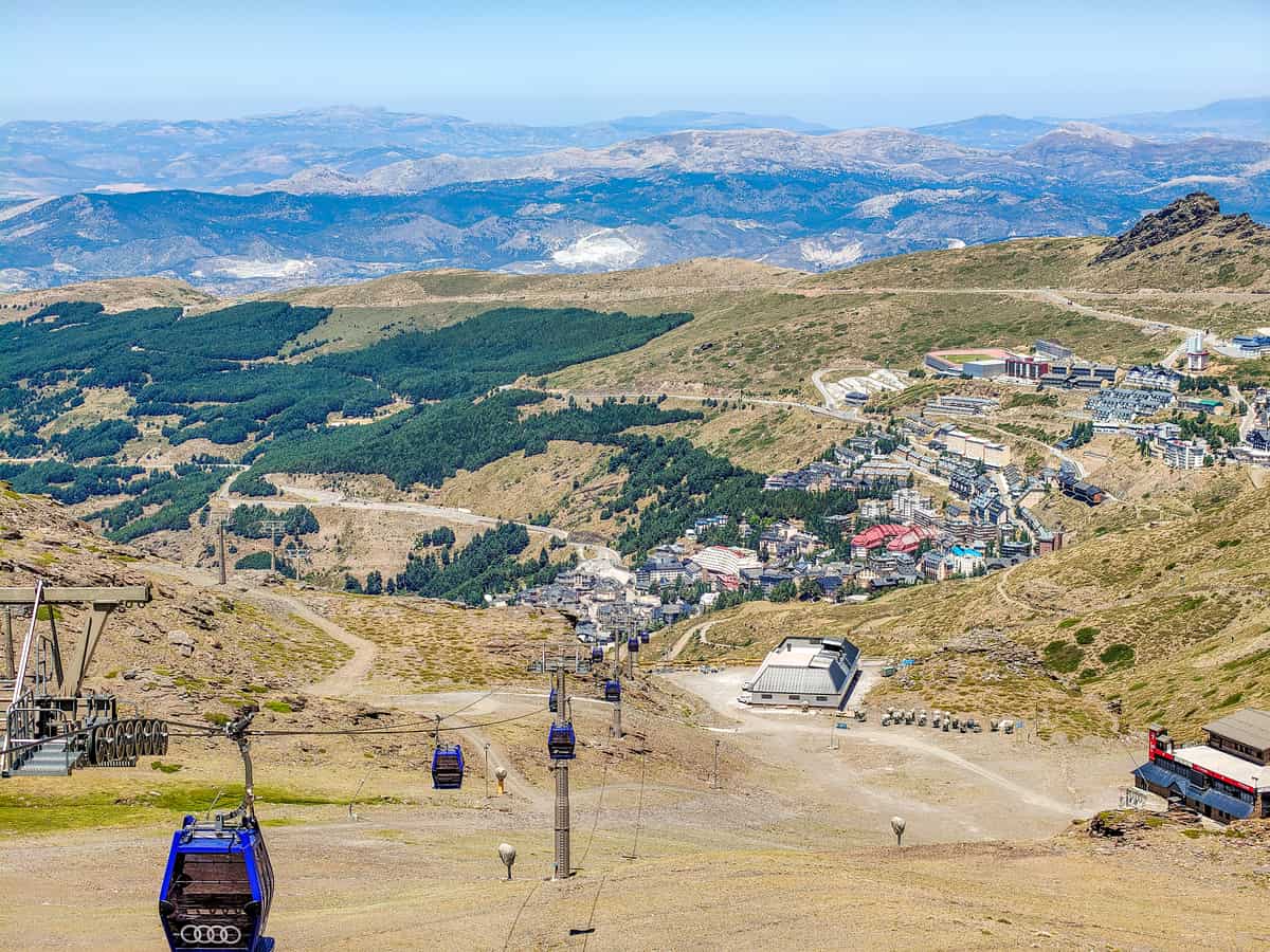 Aerial view of a mountainous landscape with cable cars travelling over hills, a small town below, green forests, and distant mountain ranges under a clear blue sky. Sierra Nevada in the summer