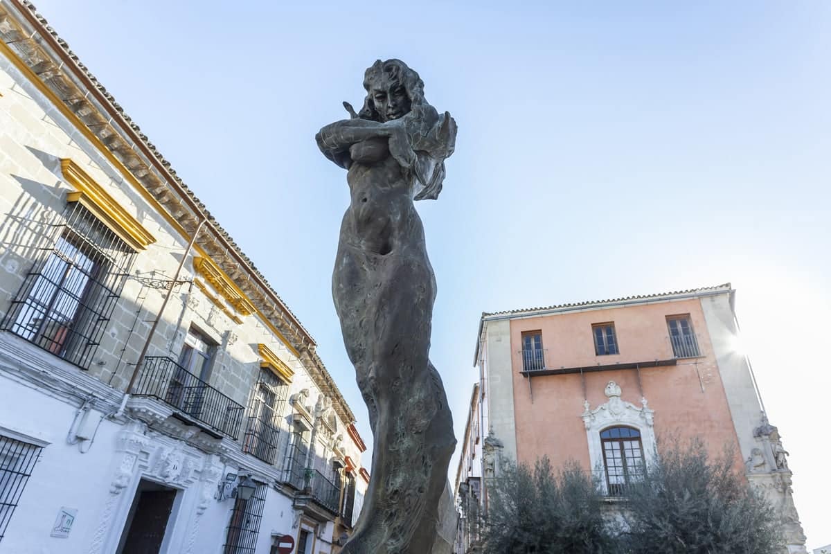 Bronze sculpture of a standing woman with flowing hair, positioned between historic buildings under a clear blue sky. Lola Flores Monument in Jerez