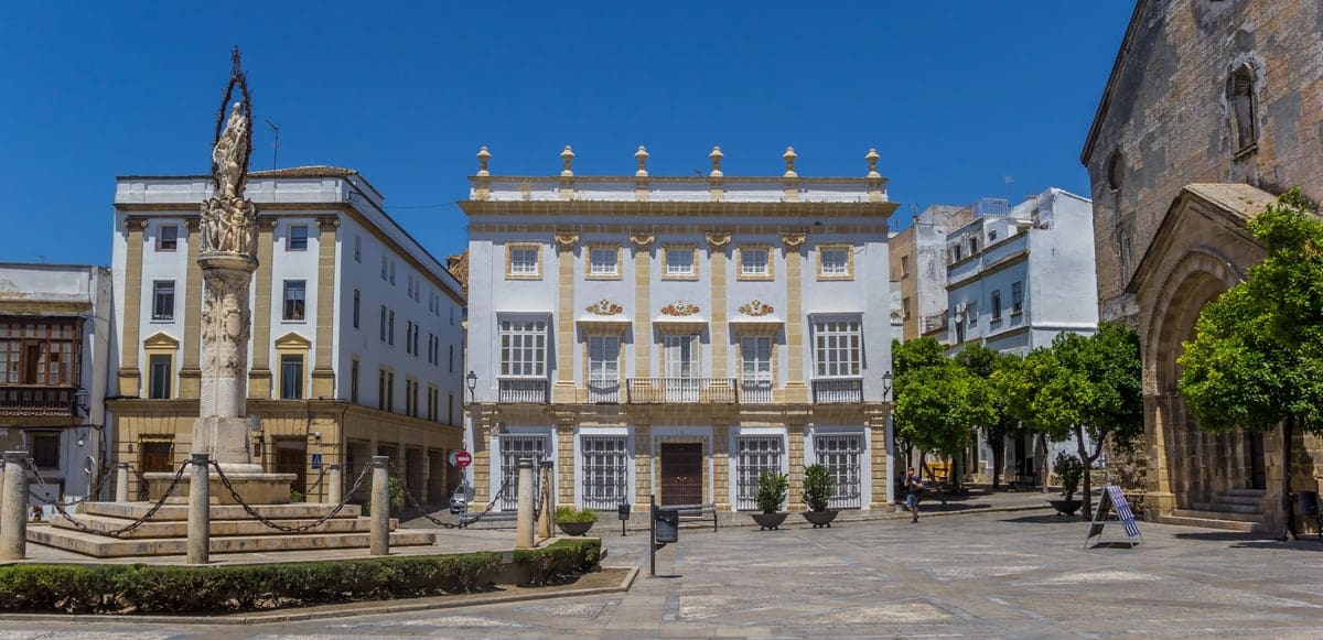 A town square featuring a stone monument, a symmetrical white building with neoclassical detailing, and an adjacent stone church. Square of of San Dionisio in Jerez