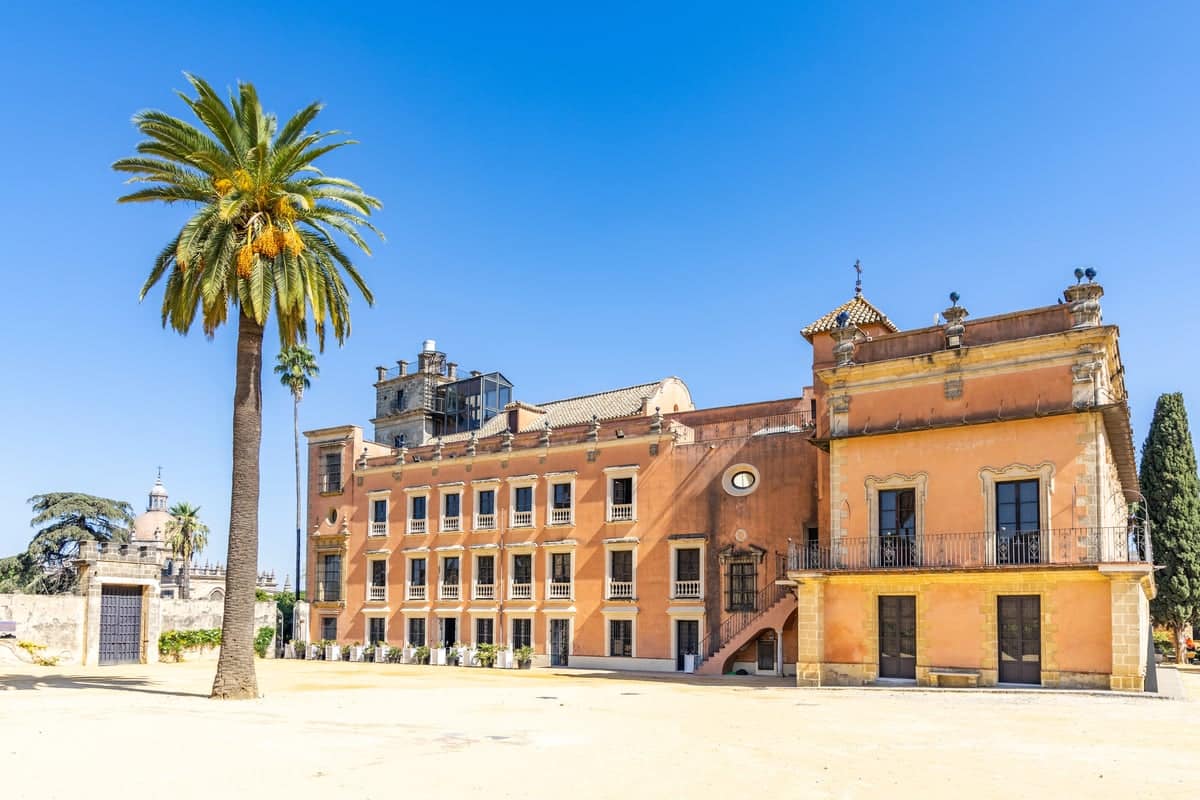 A large, multi-storey peach-coloured historic building with a balcony and many windows, located in a sandy courtyard with a tall palm tree under a clear blue sky. Villavicencio Palace in Jerez de la Frontera, Spain